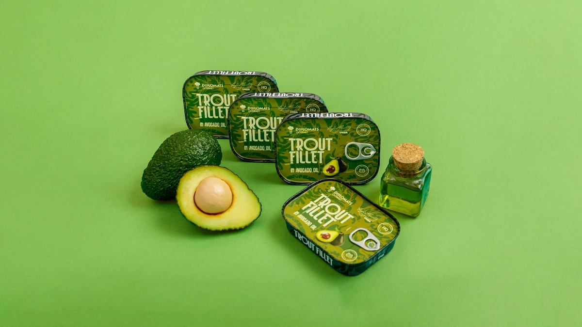 Diplomats Trout Fillet avocado oil tins beside a halved avocado on green background